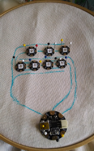 Fabric in a hoop, with a microcontroller and sevel LEDs pinned in place. Traces are drawn in fabric marker to show where conductive thread will be stitched.