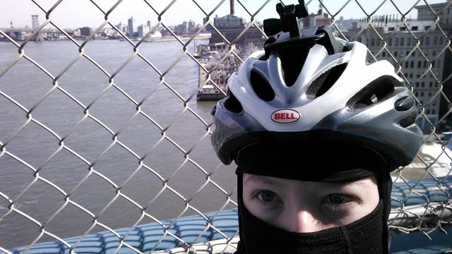 The author wearing a black balaclava under a helmet, standing before a view of the East River from the Manhattan Bridge.