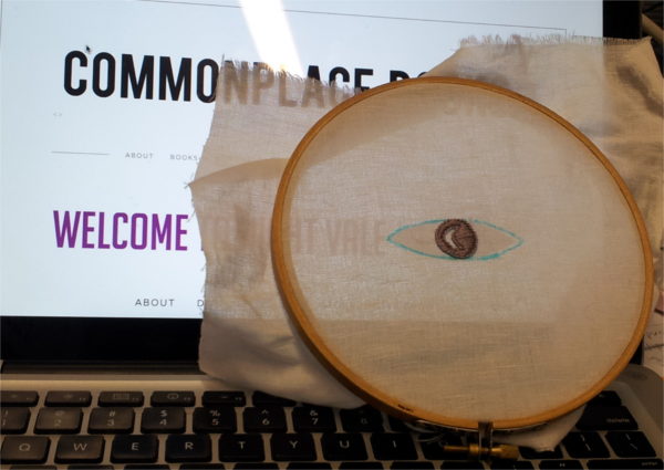 A laptop screen displays the Welcome to Night Vale banner page.  An embroidery hoop with an outline of the Night Vale logo is placed in front of the laptop screen, superimposing the embroidery over the logo.