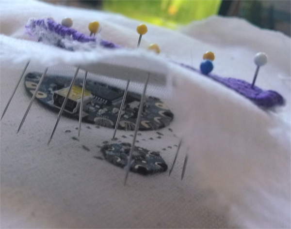 The logo is pinned in place atop a piece of linen, which is atop the canvas backing.  The light sensor and microcontroller board are visible between the intermediate linen and the canvas.