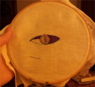 The Welcome to Night Vale logo beginning to take shape, with the iris filled in and the fill elements of the sclera approximately three-fourths complete.  The borders of the fill do not match the fabric-marker guides for the design.